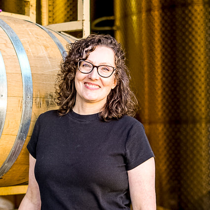 Shrisada, with glasse,s standing next to a wooden barrel in a winery setting